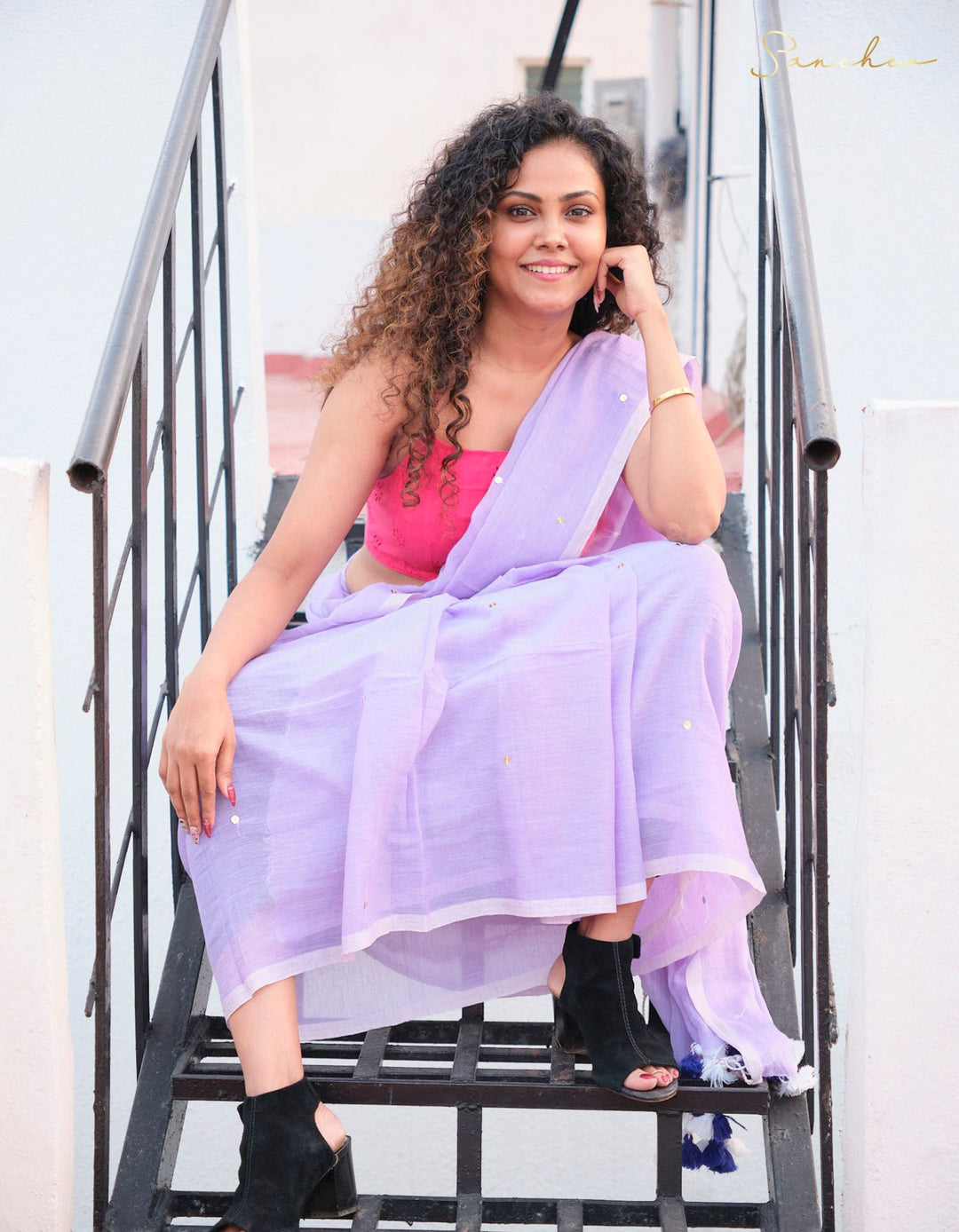 A smiling woman with curly hair sits on metal stairs, wearing a lavender mulmul cotton saree with a bright pink blouse. The saree is draped casually, showcasing its versatility for office wear. The word "Sanchen" is visible in gold script in the top right corner. Mul Cotton Sarees, Casual Sarees for Office Wear, Saree Boutique in Anna Nagar