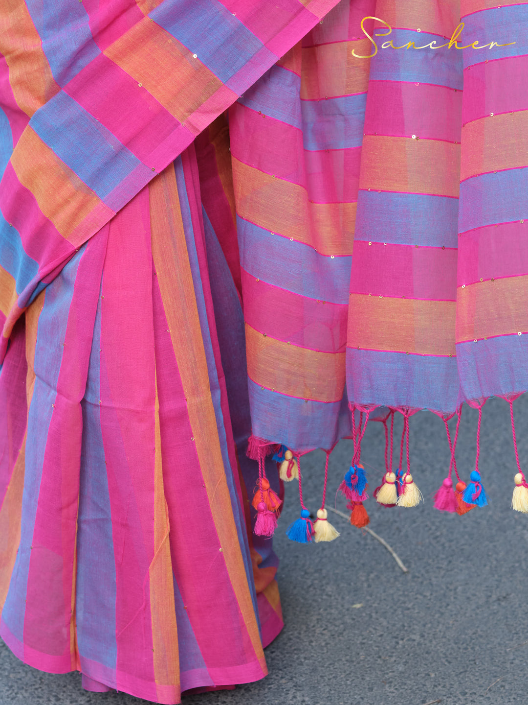 Close-up of a vibrant multicolored Mulmul cotton saree with pink, blue, and orange stripes. The saree features small tassels in various colors along the border, Mul Cotton Sarees, Workwear sarees, Saree Boutique in Anna Nagar, Best Sarees Shops in Anna Nagar, Professional Office wear Sarees, Casual Sarees for Office Wear.