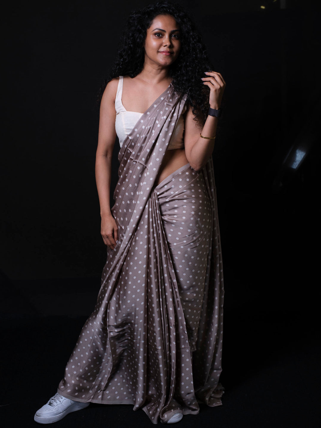Woman wearing dusty taupe modal silk saree with white polka dots and white sneakers against dark background