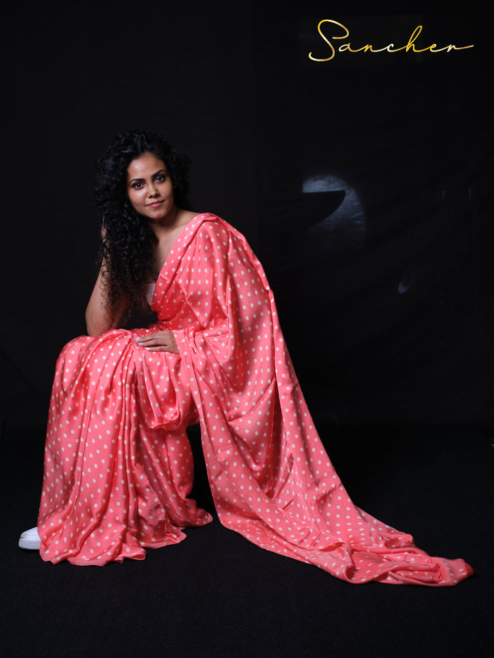 Woman sitting gracefully in coral pink polka dot modal silk saree on black background showcasing elegant draping style