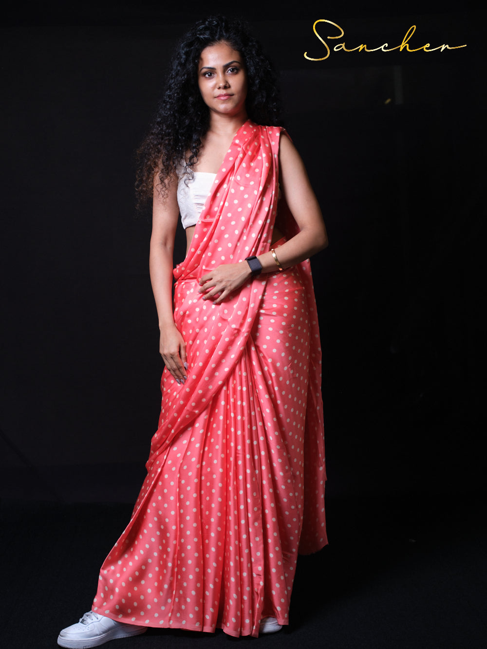Young woman standing confidently wearing coral pink polka dot saree with white sneakers for modern fusion look, perfect for parties, office occasion.