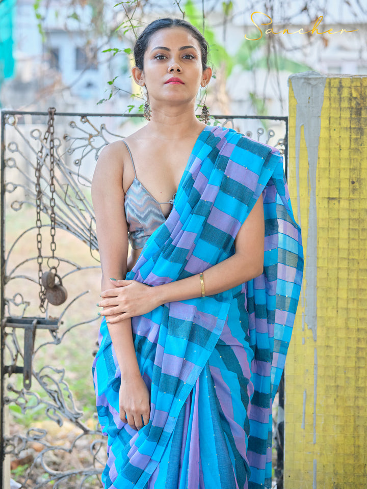 Full-length shot of woman in blue and purple striped mulmul cotton saree, standing by yellow wall and iron gate. Stylish workwear saree from top saree shops in Anna Nagar.