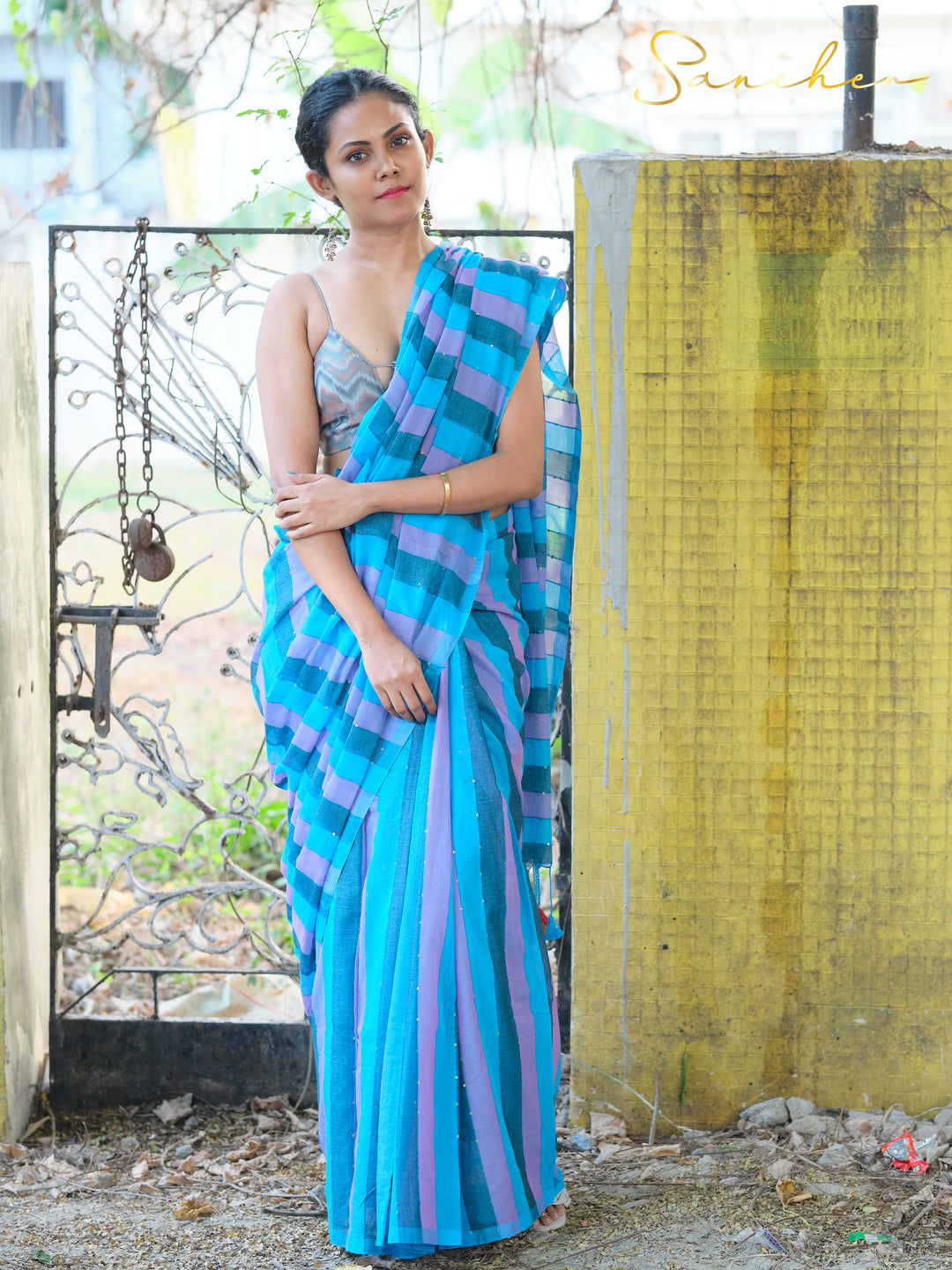 Full-length shot of woman in blue and purple striped mulmul cotton saree, standing by yellow wall and iron gate. Stylish workwear saree from top saree shops in Anna Nagar.
