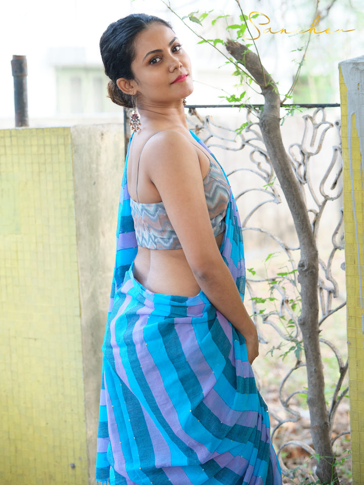 Woman in blue and purple striped mulmul cotton saree with sleeveless blouse, standing outdoors near yellow wall and plants. Perfect workwear saree from saree boutique in Anna Nagar.