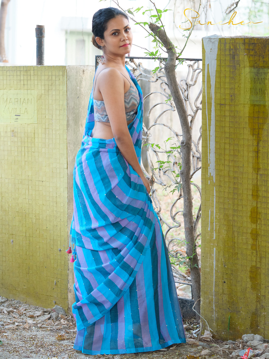 Woman in blue and purple striped mulmul cotton saree with sleeveless blouse, standing outdoors near yellow wall and plants. Perfect workwear saree from saree boutique in Anna Nagar.