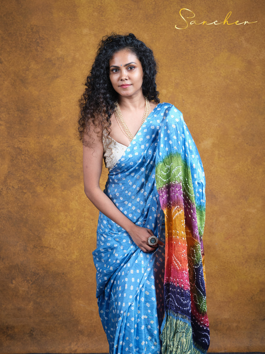 A woman wearing a vibrant blue Mulmul cotton saree with white polka dots and a colorful rainbow-hued pallu, perfect for office wear. The saree is from Sanchen, a saree boutique in Anna Nagar known for professional workwear sarees.
