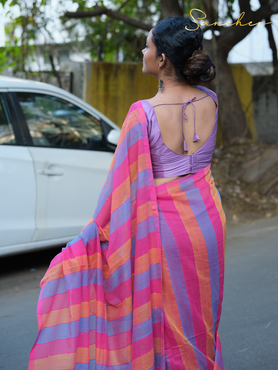 Back view of a woman wearing a striped Mulmul cotton saree in pink, blue, and orange colors. The saree is paired with a purple blouse featuring an open back design with tie closures, Mul Cotton Sarees, Workwear sarees, Saree Boutique in Anna Nagar, Best Sarees Shops in Anna Nagar, Professional Office wear Sarees, Casual Sarees for Office Wear