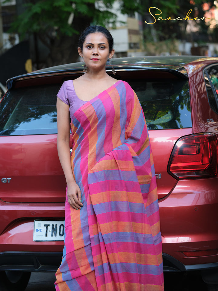 ront view of a woman wearing a colorful striped Mulmul cotton saree in pink, blue, and orange. She stands in front of a red car, showcasing the saree as casual office wear, Mul Cotton Sarees, Workwear sarees, Saree Boutique in Anna Nagar, Best Sarees Shops in Anna Nagar, Professional Office wear Sarees, Casual Sarees for Office Wear