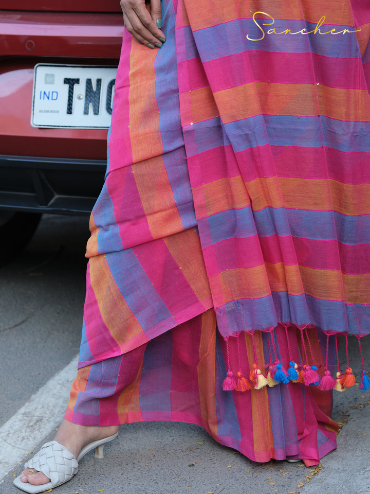 A woman wearing a colorful striped Mulmul cotton saree stands next to a car with an Indian license plate. The saree has pink, blue, and orange stripes with colorful tassels at the hem, Mul Cotton Sarees, Workwear sarees, Saree Boutique in Anna Nagar, Best Sarees Shops in Anna Nagar, Professional Office wear Sarees, Casual Sarees for Office Wear.