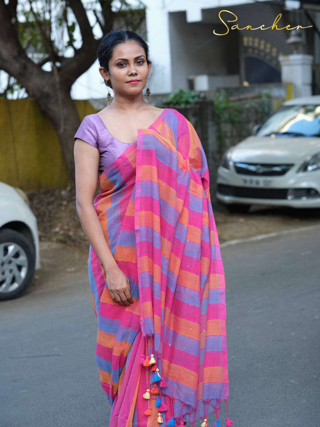 A woman models a bright striped Mulmul cotton saree in pink, blue, and orange hues. She wears a purple blouse and stands in an outdoor setting with cars and trees in the background, Mul Cotton Sarees, Workwear sarees, Saree Boutique in Anna Nagar, Best Sarees Shops in Anna Nagar, Professional Office wear Sarees, Casual Sarees for Office Wear.