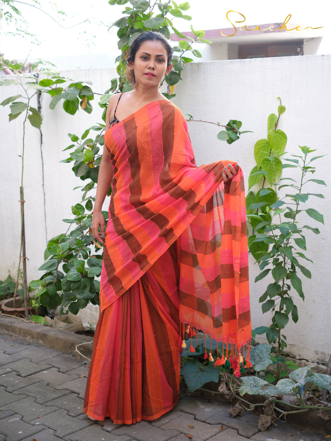 Woman wearing vibrant orange and pink striped mul cotton saree in garden setting, featuring casual sarees for office wear from Sancher, best saree shop in Anna Nagar.