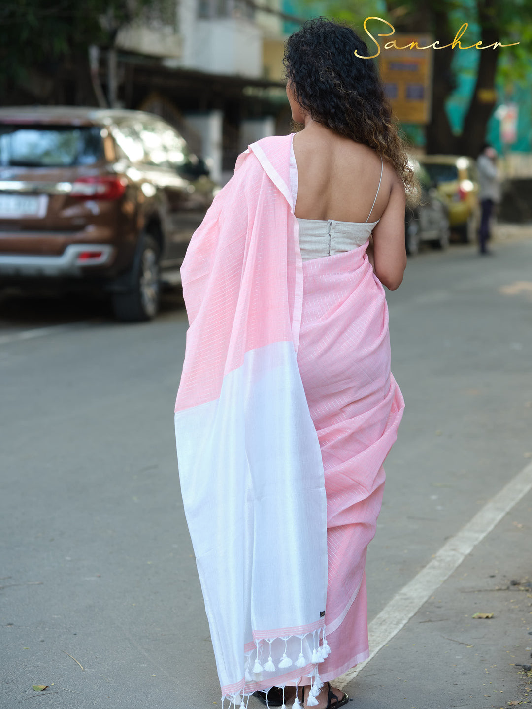 Woman wearing a pink and white mul cotton saree with tassels, viewed from behind on a city street, showcasing its suitability for office wear. Keywords: Mul Cotton Sarees, Workwear sarees, Casual Sarees for Office Wear, Pink sarees Online